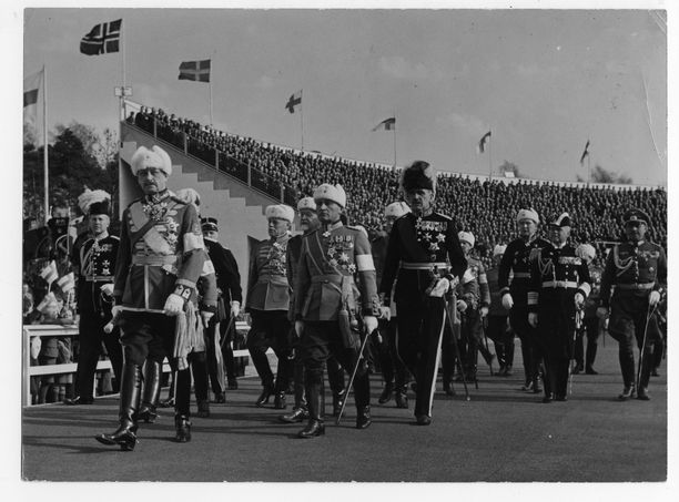 Mannerheim seurueineen saapuu vasta valmistuneelle Helsingin olympiastadionille puolustusvoimain lippujuhlanpäivän paraatiin 16. toukokuuta 1938. Valkoiset hihanauhat näkyvät käsivarsissa.