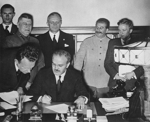 Diktaattori Josif Stalin myhäilee takana, kun ulkoministerit Molotov ja Ribbentrop allekirjoittajat sopimusta Euroopan jaosta.